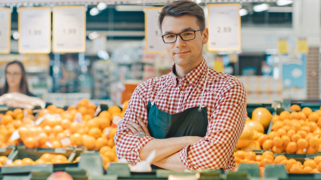 A worker smiling in front of the fruit section of the supermarket