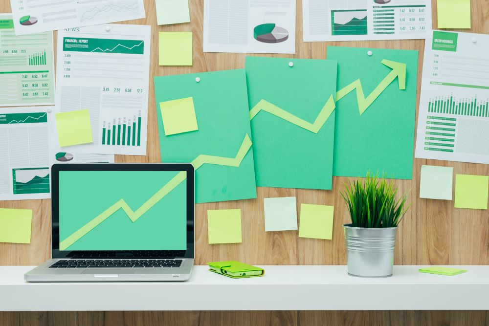 The image depicts a workspace featuring a laptop with a green upward-trending graph displayed on the screen. The background includes several charts and graphs pinned to a wooden wall, all showing positive trends. Additionally, there are numerous yellow sticky notes attached to the wall. On the desk, there is a small potted plant and a green smartphone next to the laptop.
