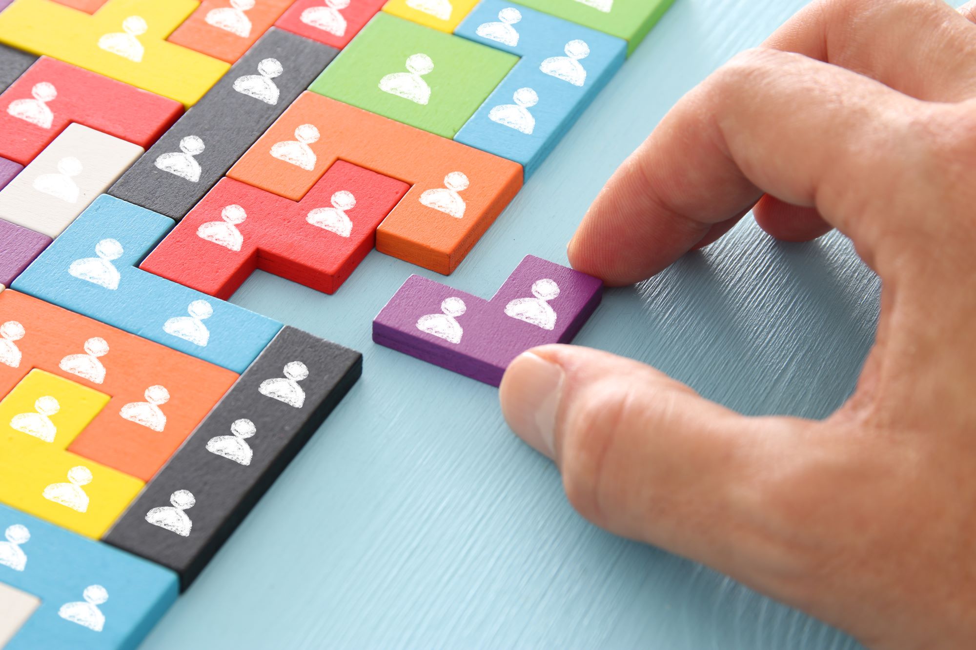 The image shows a hand placing a purple puzzle piece with a white human icon on it among other colorful puzzle pieces. Each puzzle piece has a white human icon, and the pieces are in various colors including red, green, blue, yellow, orange, black, and white. The background surface is light blue.