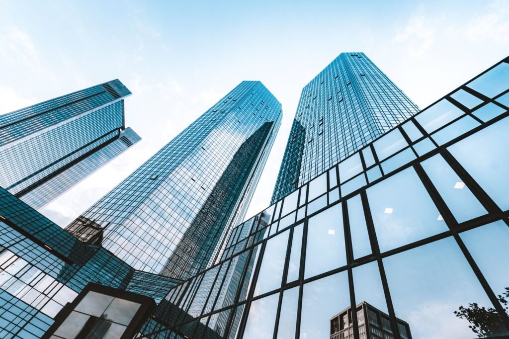 The image shows a low-angle view of several tall, modern skyscrapers with glass facades reflecting the sky and surrounding buildings. The buildings are set against a clear blue sky, and the perspective emphasizes their height and sleek architectural design.