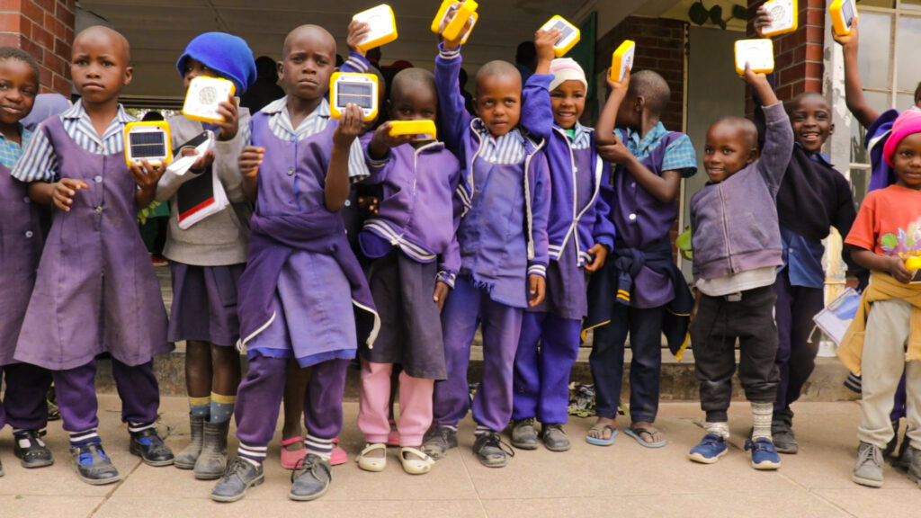 The image shows a group of children standing in a line, holding up solar buddy devices. The children are wearing uniforms consisting of purple and blue clothing. Their faces are obscured for privacy.