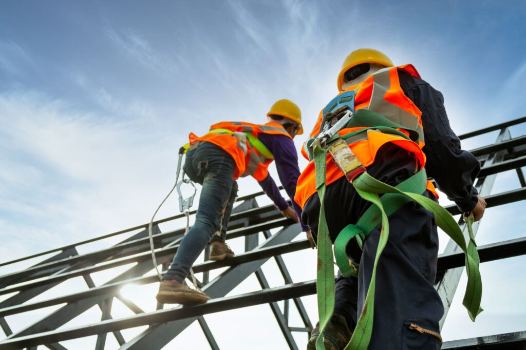 The image shows two construction workers wearing safety harnesses, helmets, and high-visibility vests. They are climbing a metal structure, possibly a roof or scaffolding, under a clear blue sky with the sun shining brightly.