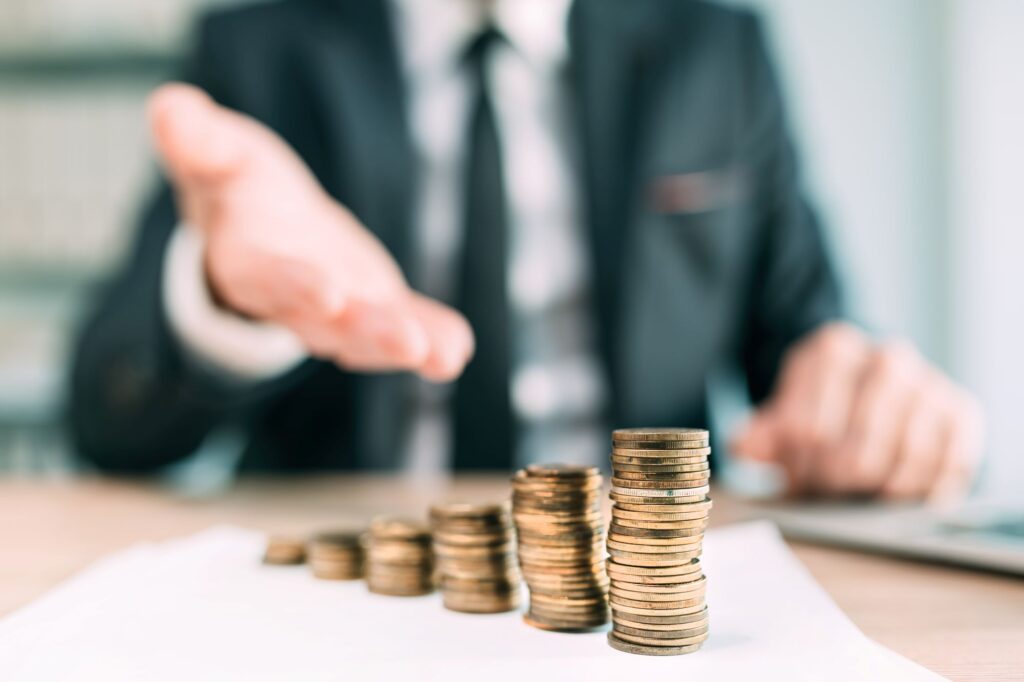 The image shows a person in a suit sitting at a desk with stacks of coins arranged in ascending order on a piece of paper. The person's hand is extended, possibly gesturing towards the coins. The background is blurred, drawing attention to the coins and the person's hand.