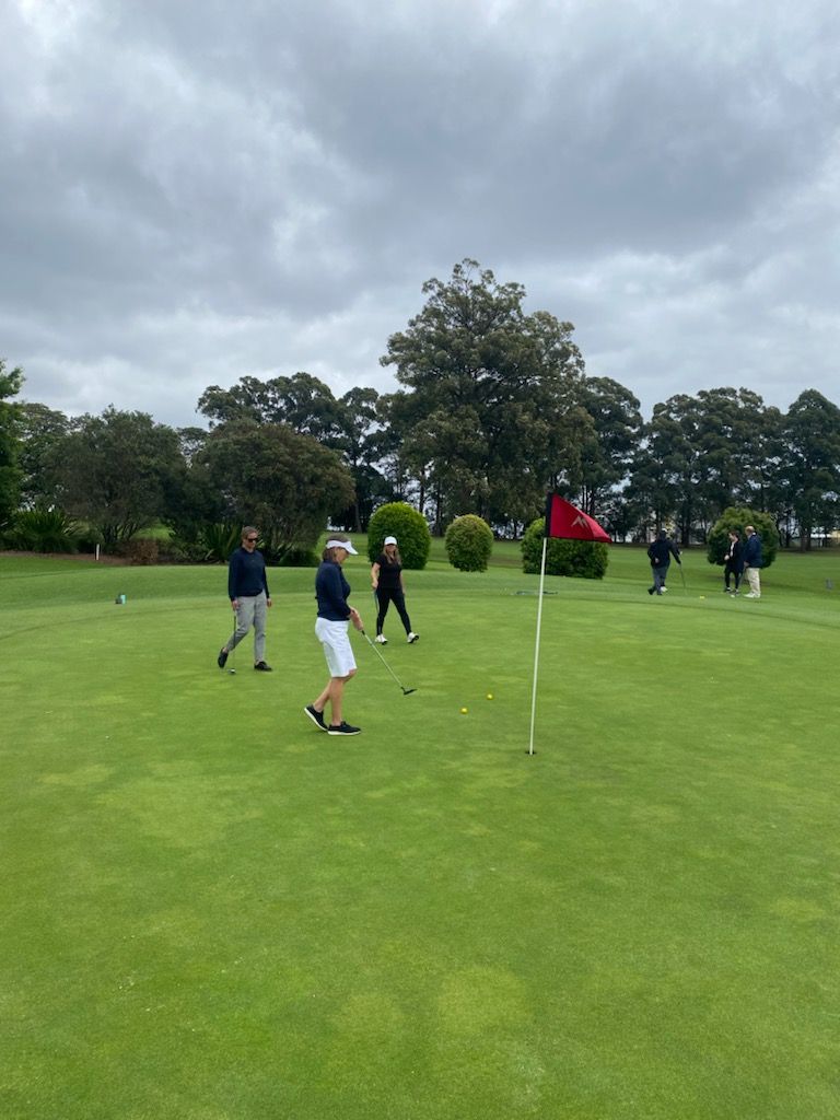 The image shows a group of people on a golf course. Three individuals are visible near the putting green, with one person preparing to putt the golf ball towards the hole marked by a red flag. The background features lush greenery, including trees and bushes, under an overcast sky.