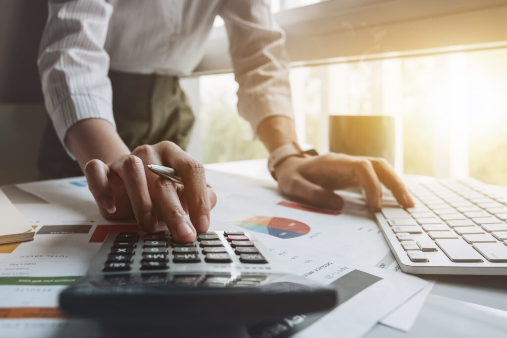 Close up of businessman or accountant hand holding pen working on calculator to calculate business data, accountancy document and laptop computer at office, business concept