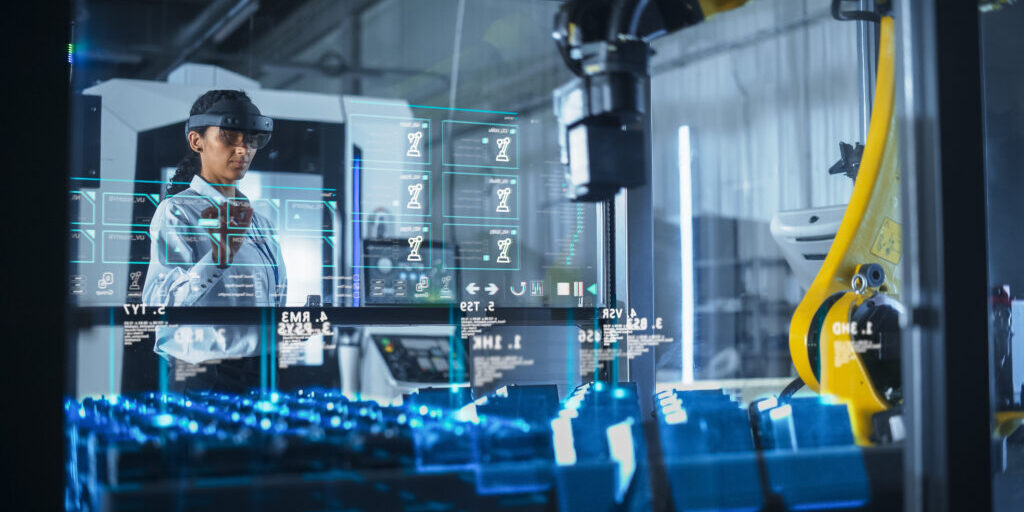 Engineer Wearing Augmented Reality Headset Operates Robotic Arm in a Factory, Interacting With Virtual Controls on a Transparent Holographic Interface. Advanced Machinery and Digital Displays.