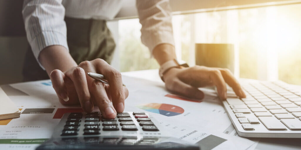 Close up of businessman or accountant hand holding pen working on calculator to calculate business data, accountancy document and laptop computer at office, business concept