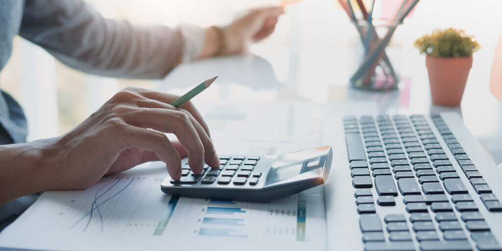 Close up of businessman or accountant hand holding pencil working on calculator to calculate financial data report, accountancy document and laptop computer at office, business concept