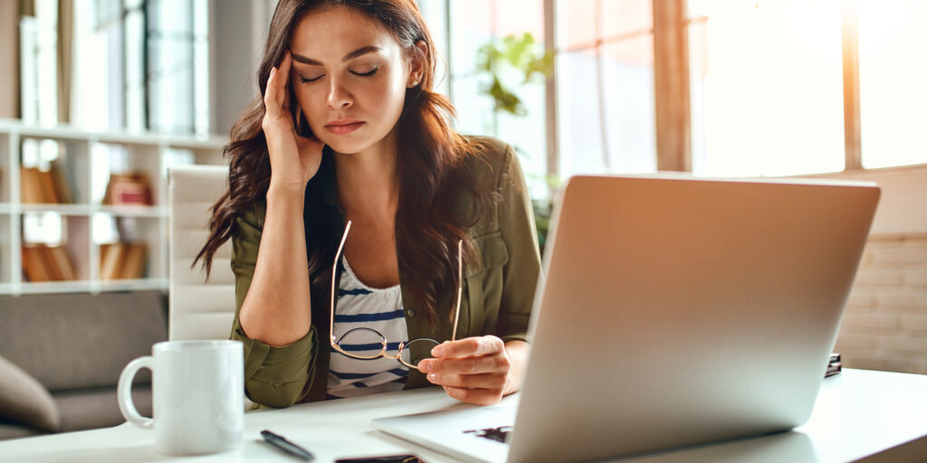 Tired business woman in stress works at a laptop while sitting at a table at home and holds her hand on her temples, migraine attack. Freelance, work from home.