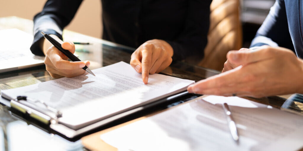 Close-up Of Two Businesspeople Hand Working On Contract Paper Over Desk